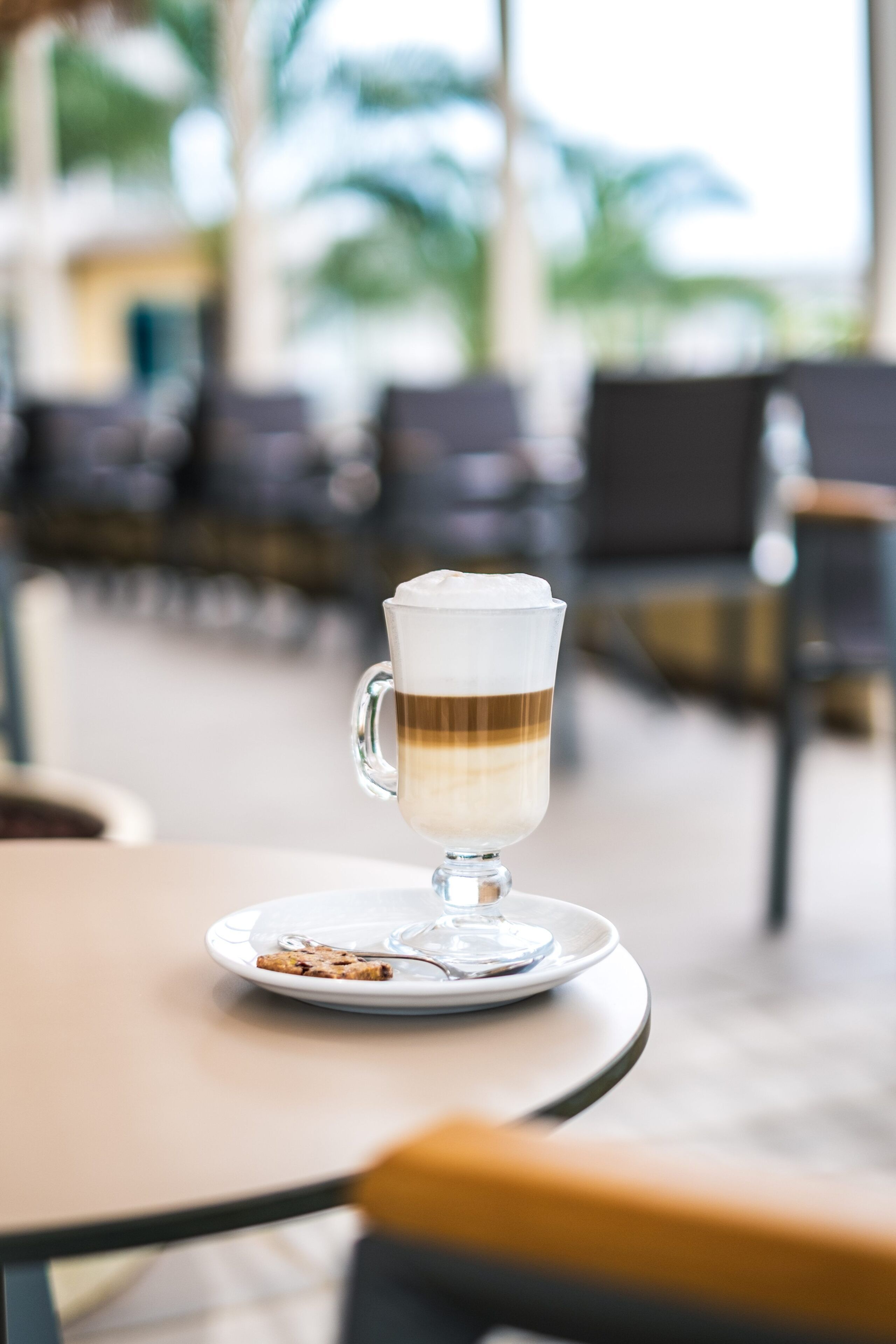 Un verre transparent contenant une boisson à plusieurs couches, posé sur une soucoupe, sur une table dans un intérieur bien éclairé, avec des chaises et un arrière-plan flou.