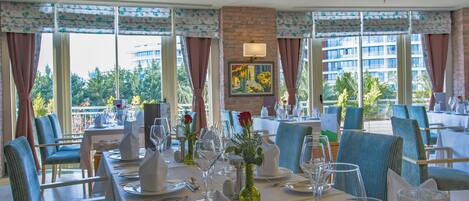 A dining area with tables set for a meal, large windows with curtains, and a brick wall with a painting.
