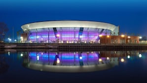 Un bâtiment moderne de forme circulaire, avec un bassin d’eau dont la surface réfléchit la lumière, situé à l’avant.
