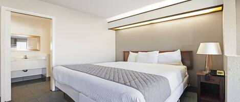 A hotel room with a large bed, a bedside table with a lamp, a mirror, and a bathroom with a sink and toilet.