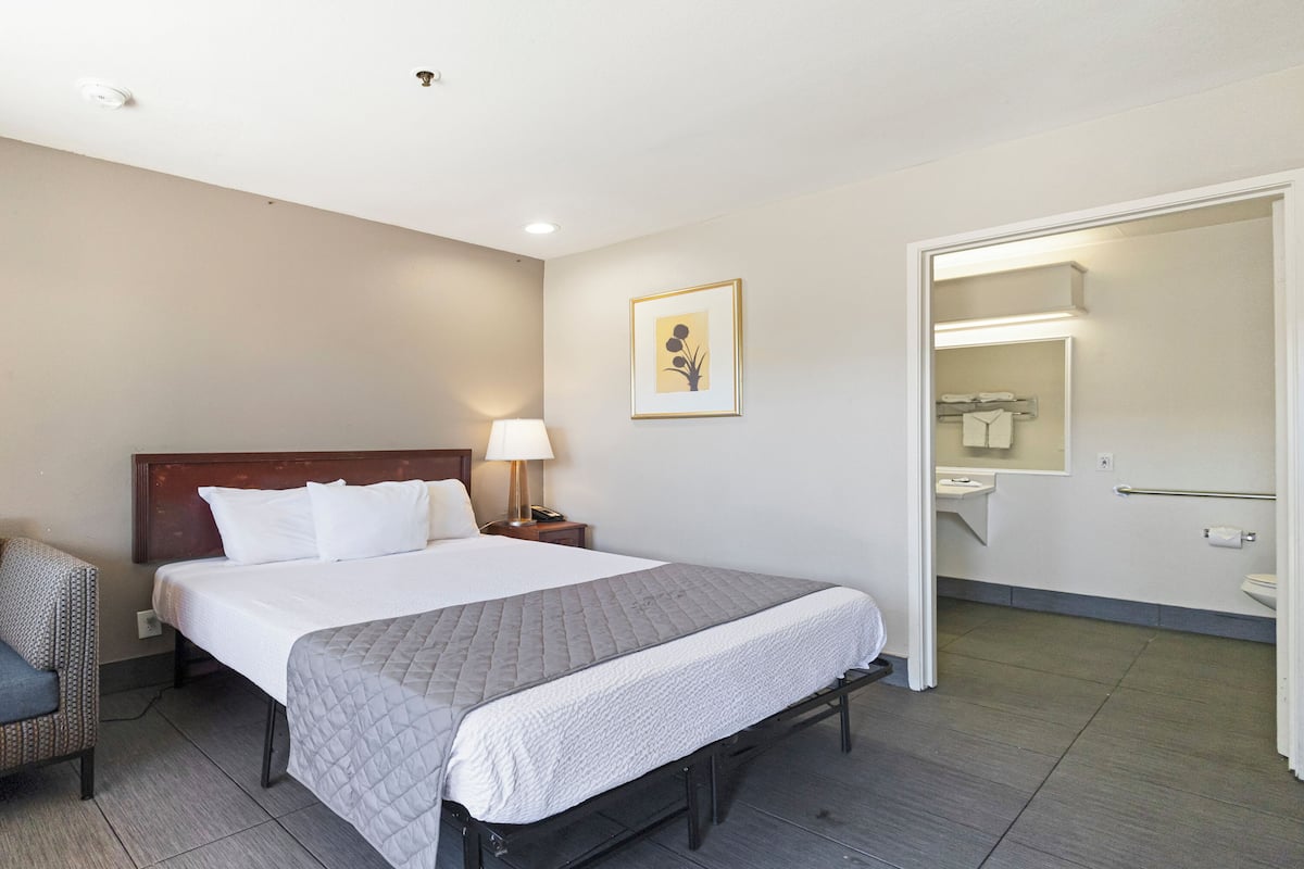 A hotel room with a bed, a chair, a bedside table with a lamp, a framed picture on the wall, and an open door leading to a bathroom.