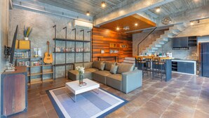 A modern living space with a sofa, coffee table, bookshelves, a guitar, and a kitchen area with a staircase. - Aonang Paradise Resort (Krabi)
