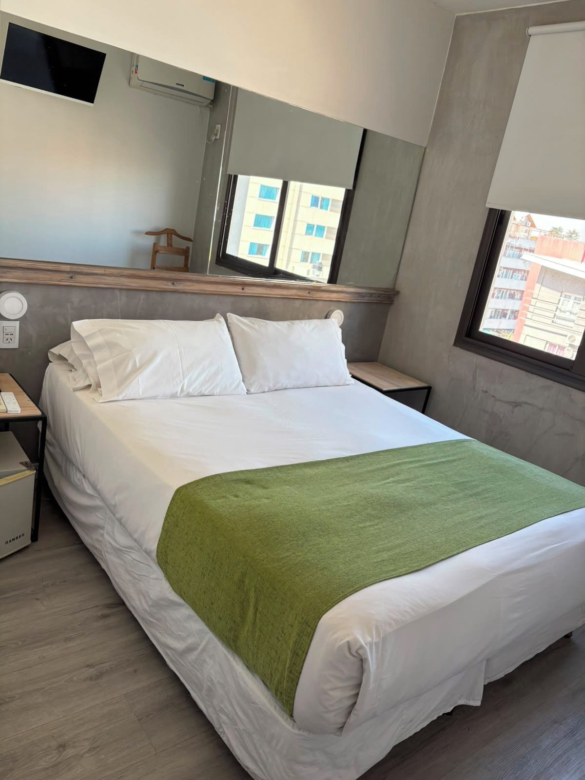 Una habitación de hotel con una cama, una mesita de noche, un escritorio con una computadora, una silla y una ventana con vistas a edificios.