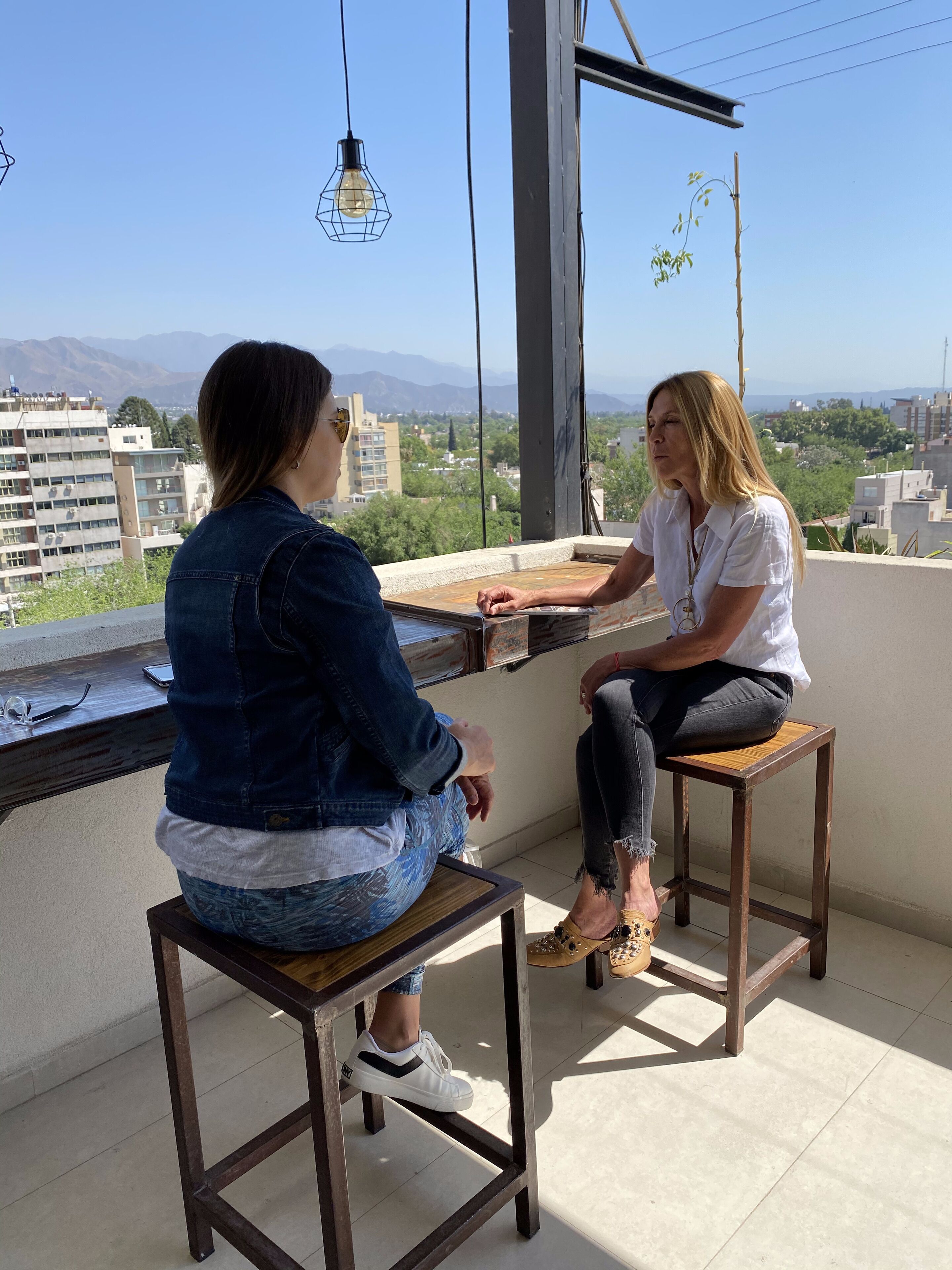 Two people sitting on wooden stools on a balcony with a city view.