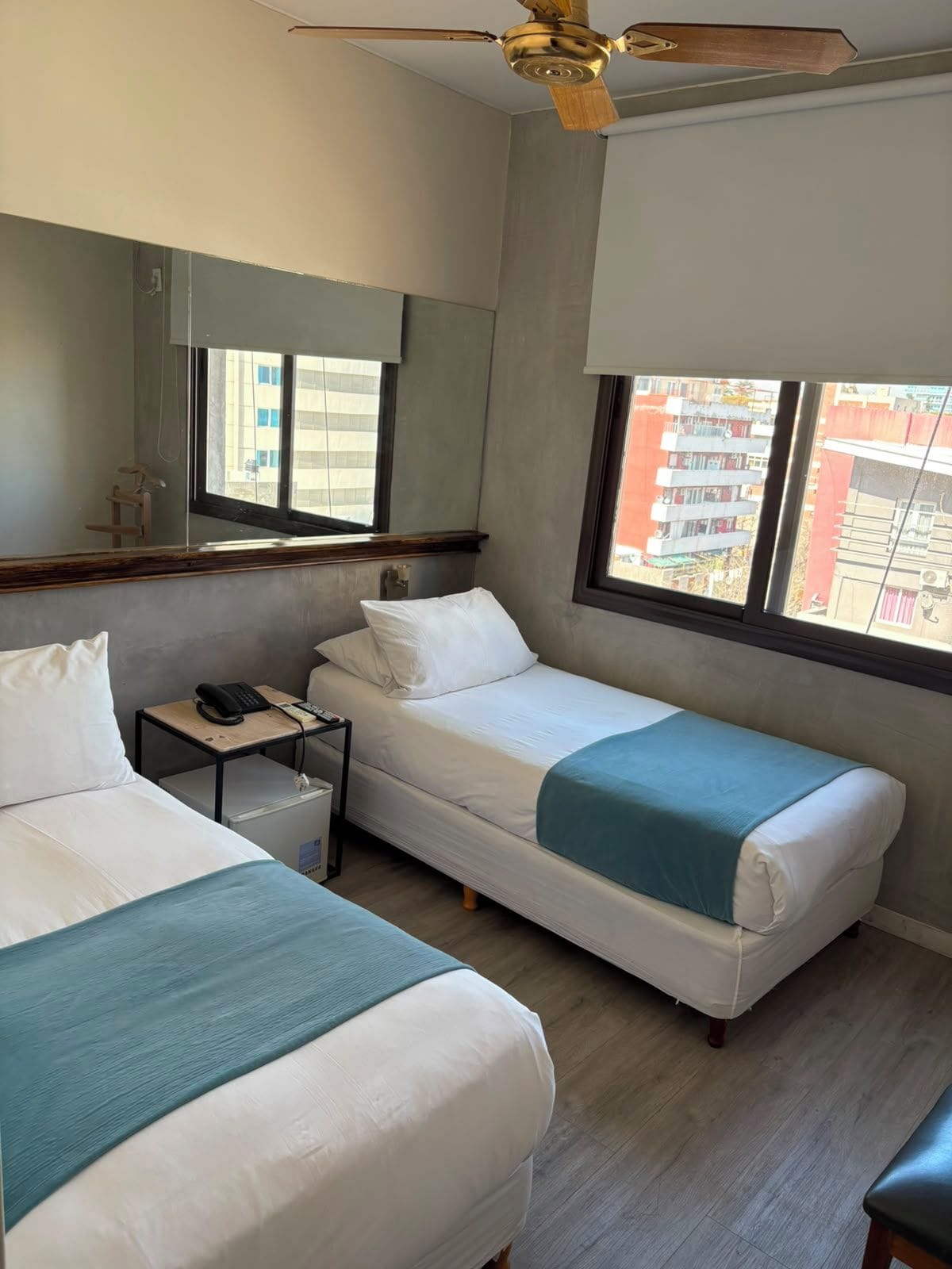 A hotel room with two beds, a ceiling fan, a small table with a phone, and a window with a view of buildings.