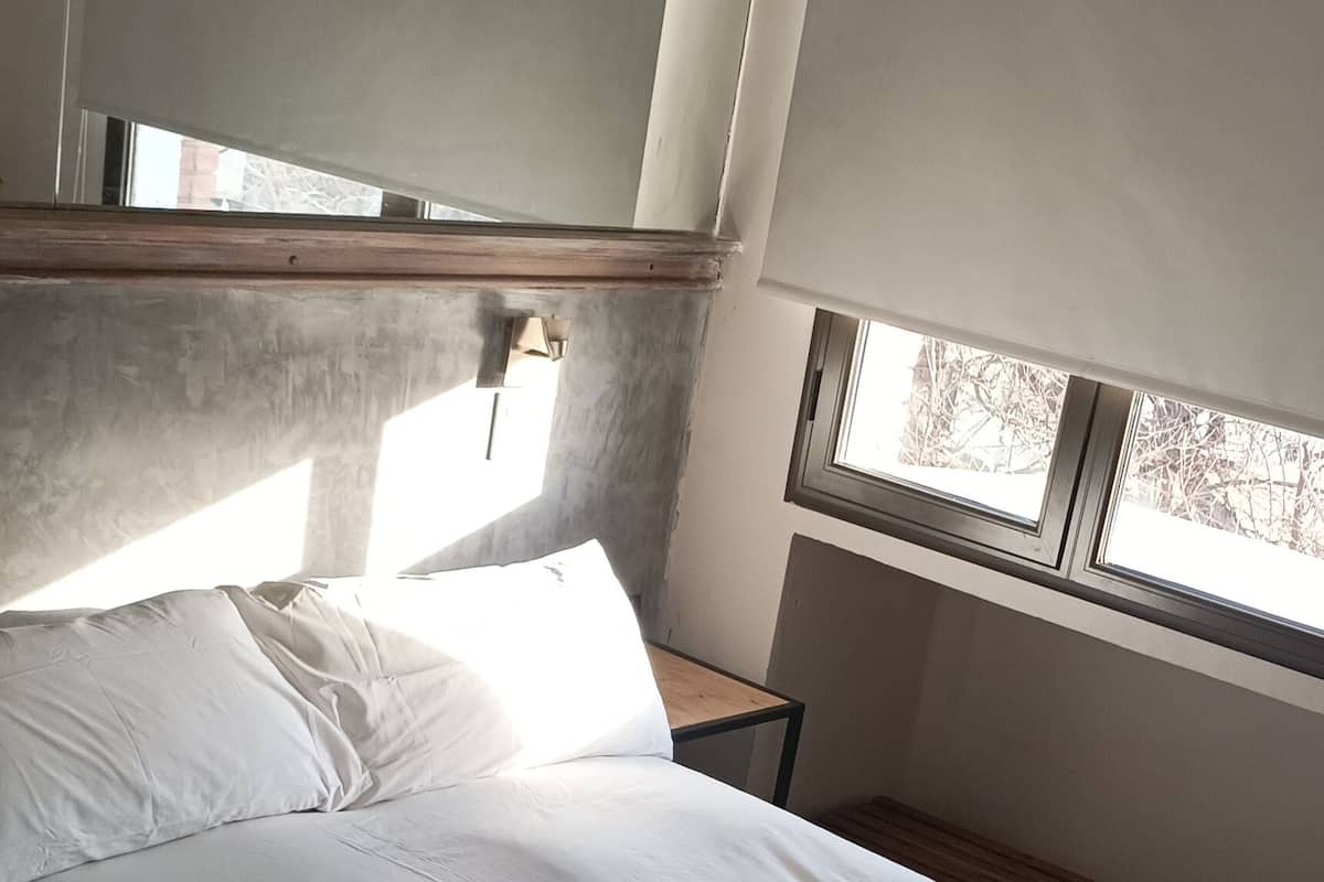 A bedroom with a bed, a blue blanket, a wooden headboard, a nightstand, and a window with a view of trees.