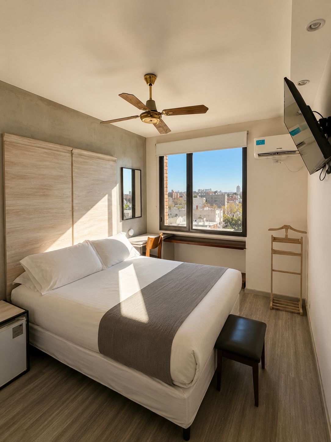 A modern hotel room with a large bed, a desk, a chair, a ceiling fan, a TV, and a window with a city view.
