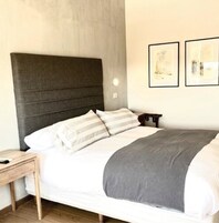 Una cama bien hecha con cabecera gris, ropa de cama blanca y una manta gris. Hay dos cuadros enmarcados en la pared sobre la cama.