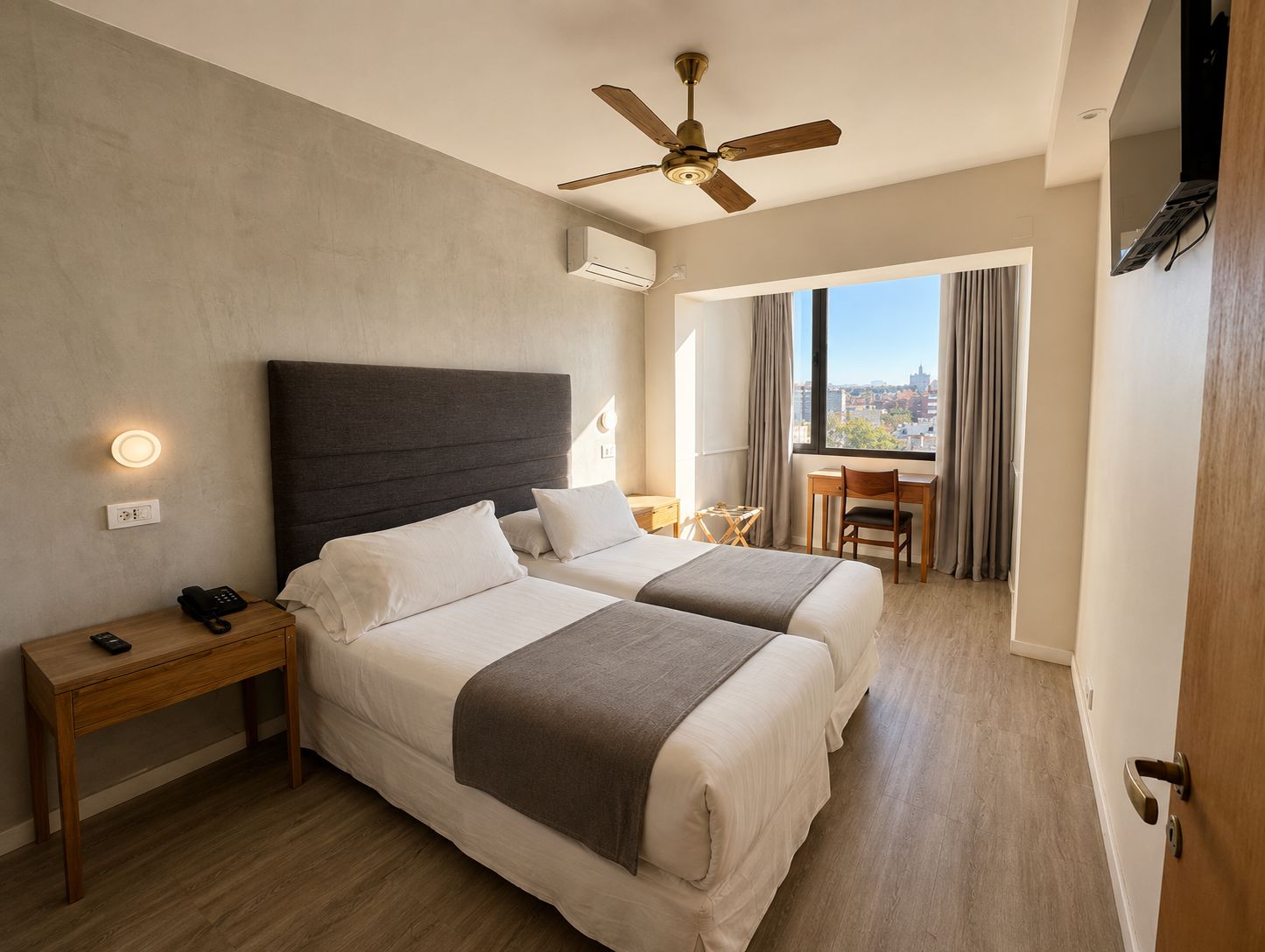 A hotel room with two beds, a desk, a chair, a ceiling fan, and a view of the city.