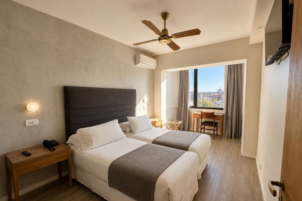 A hotel room with two beds, a desk, a chair, a ceiling fan, and a view of the city.