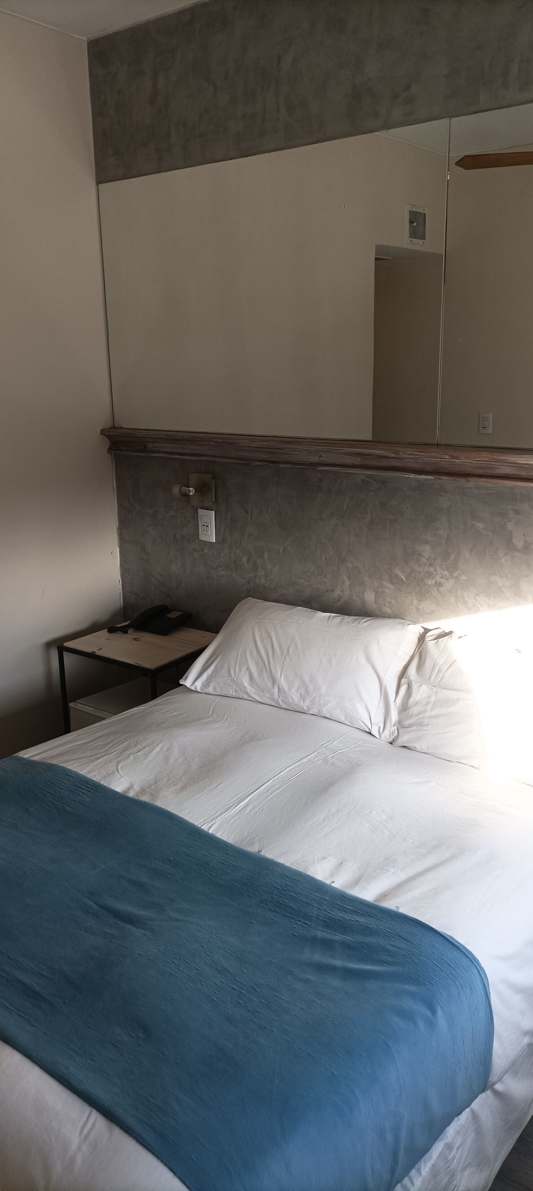 Een bed met witte lakens en een blauwe deken, een nachtkastje met een telefoon, en een spiegel aan de muur.