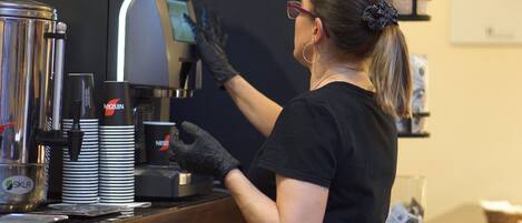 Een persoon bedient een koffieapparaat achter een toonbank, met gestapelde bekers en een koffiekan met de tekst 'Nescafe'.
