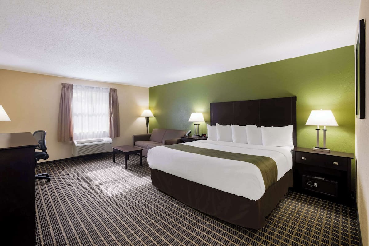 A hotel room with a large bed, two bedside tables with lamps, a desk with a chair, a sofa, and a window with curtains.