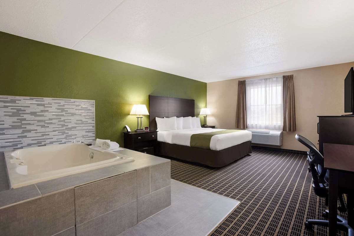 A hotel room with a large bathtub, a bed with a dark headboard, two bedside lamps, a window with curtains, and a flat-screen TV.