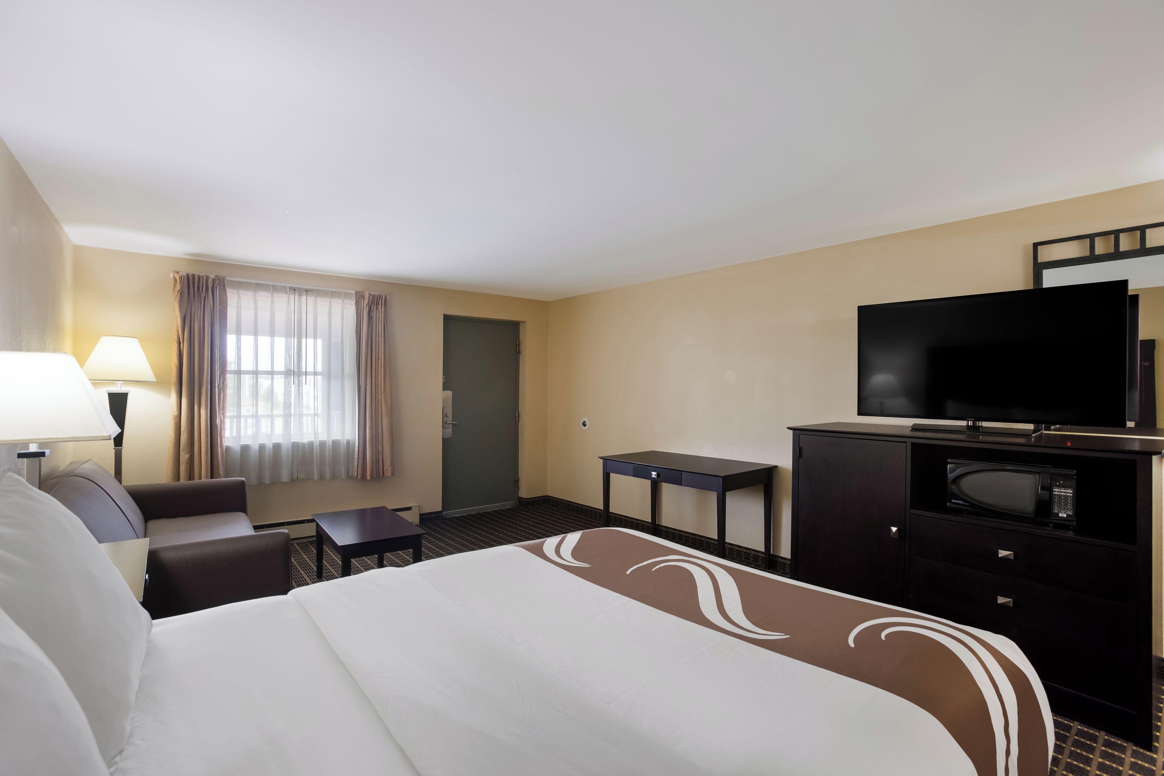 A hotel room with a bed, a desk, a television, a microwave, and a window with curtains.