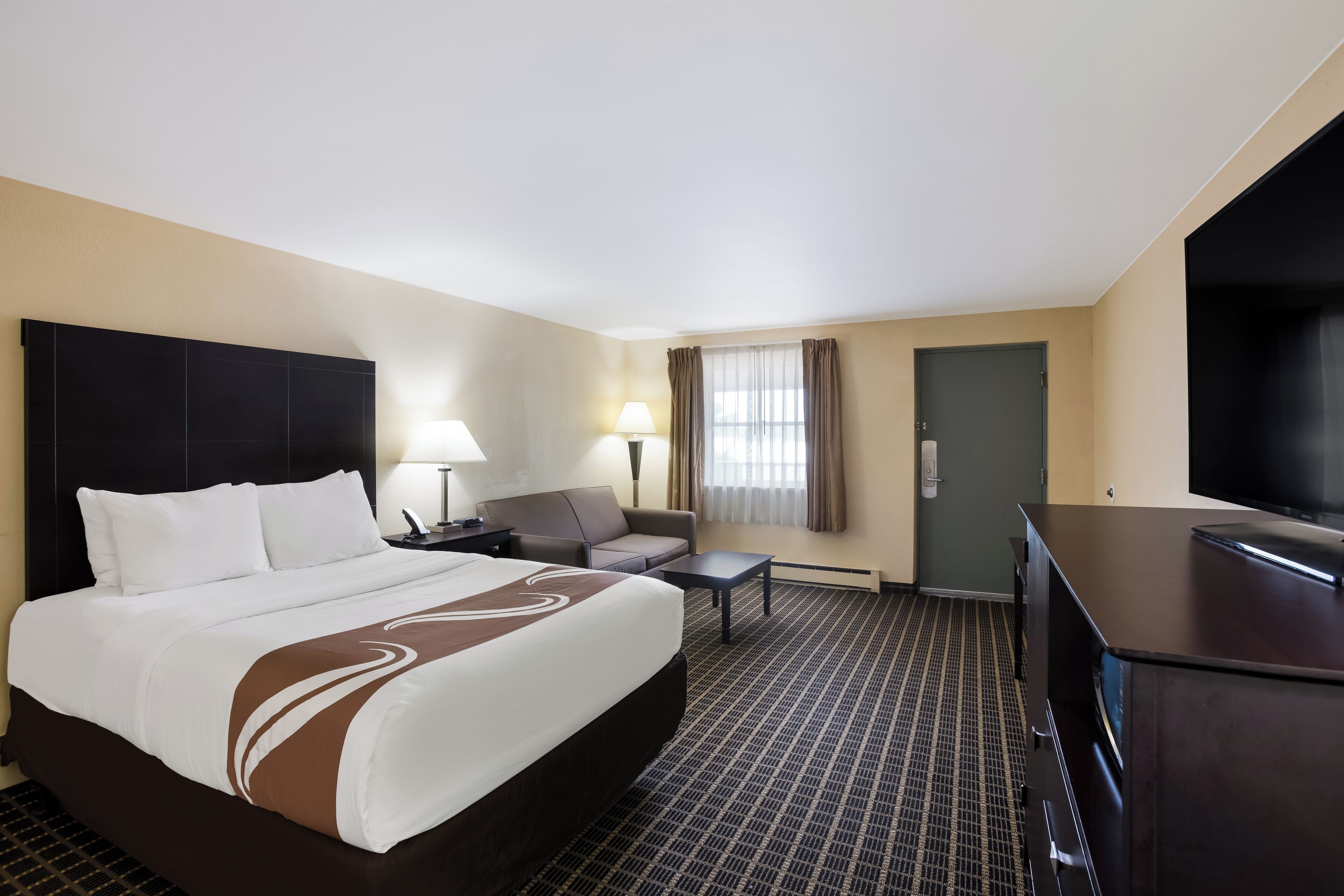 A hotel room with a large bed, a sofa, a desk, a TV, and a door.