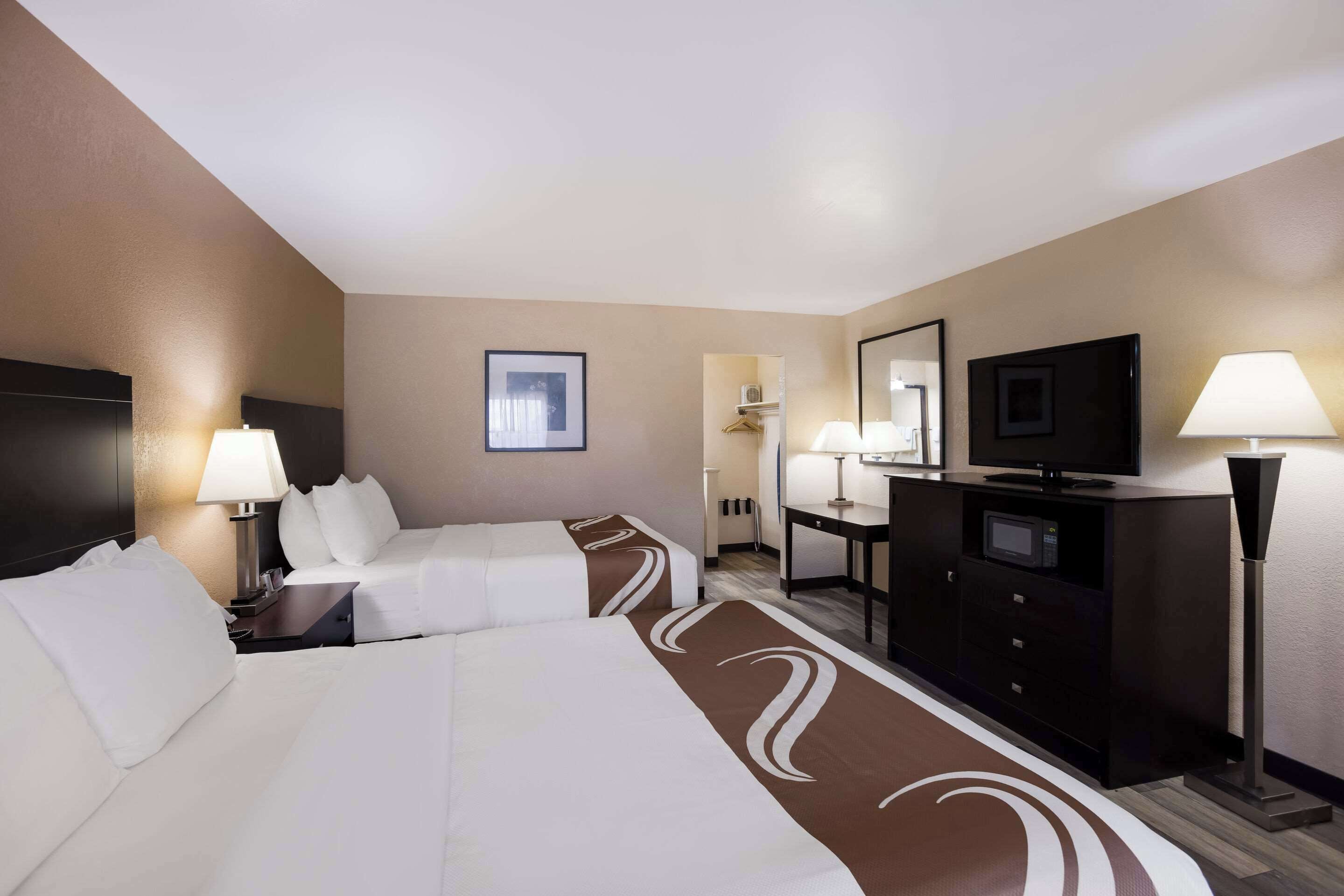 A hotel room with two beds, a desk, a TV, and a lamp.