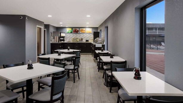 Un restaurant moderne avec des tables et des chaises blanches, un espace cuisine à l’arrière-plan et une vue sur la rue grâce à de grandes fenêtres.