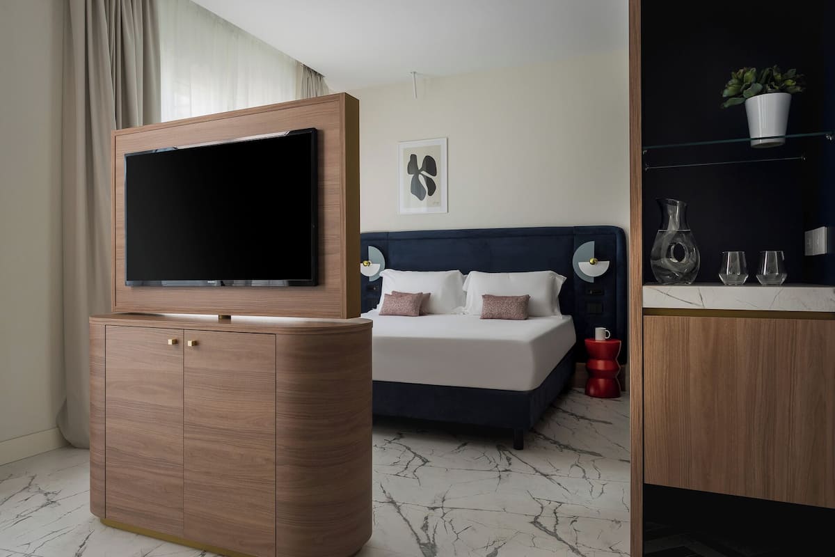 A modern hotel room with a flat-screen TV mounted on a wooden cabinet, a bed with a dark headboard, a bedside table with a lamp, and a small shelf with decorative items.