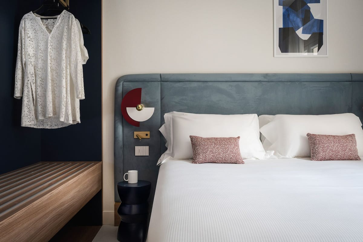 A modern hotel room with a large bed, a nightstand, a wall-mounted clock, and a hanging garment.
