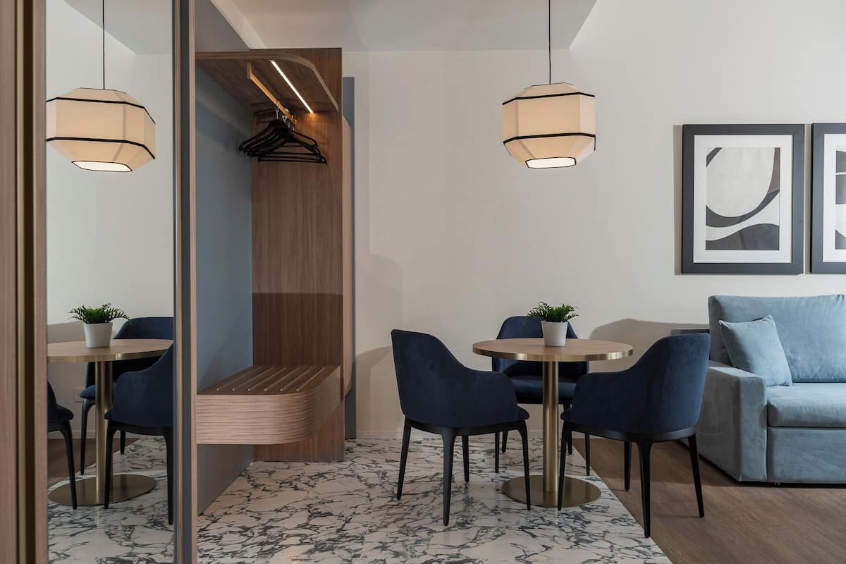 A modern hotel room with a seating area, a dining table, and a sofa. There are two pendant lights and a wall-mounted lamp.