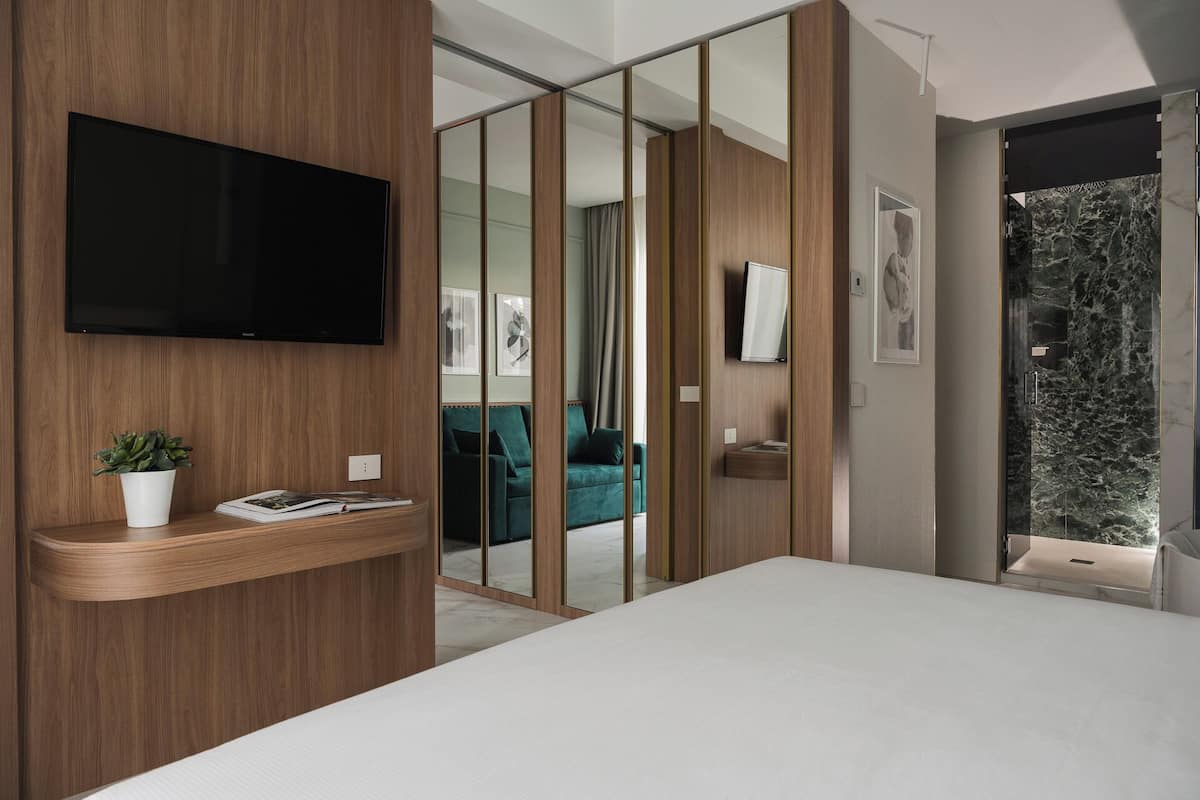 A modern bedroom with a large bed, a TV mounted on a wooden panel, a small plant on a bedside table, and a sliding glass door leading to a living area with a sofa and curtains.