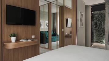 Une chambre moderne avec un grand lit, un téléviseur fixé sur un panneau en bois, une petite plante sur une table de chevet et une porte-fenêtre coulissante donnant sur un espace de vie avec un canapé et des rideaux.