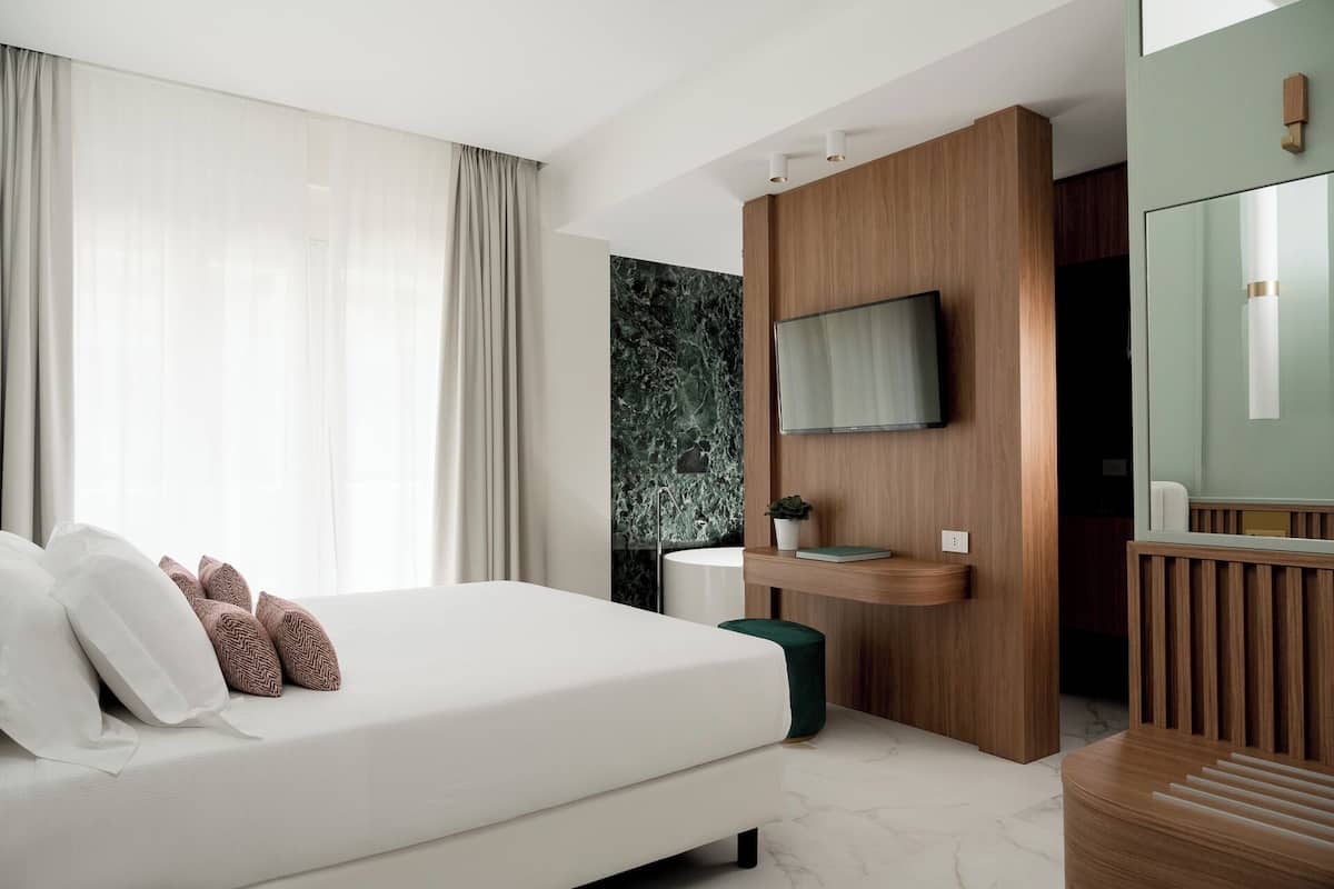 A modern hotel room with a large bed, a mounted TV, a wooden vanity, and a marble wall.