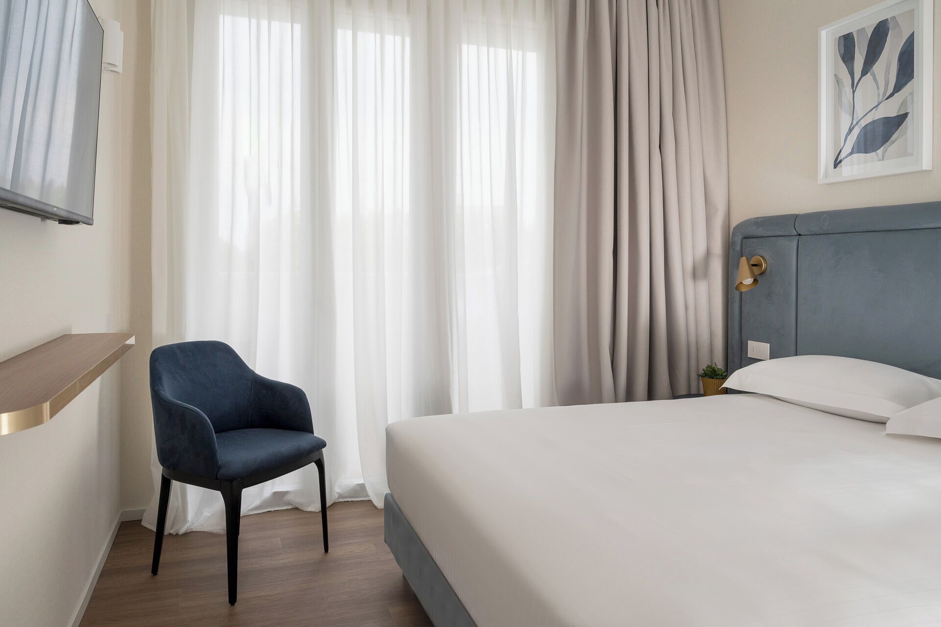 A hotel room with a bed, a chair, a TV, and a painting on the wall.