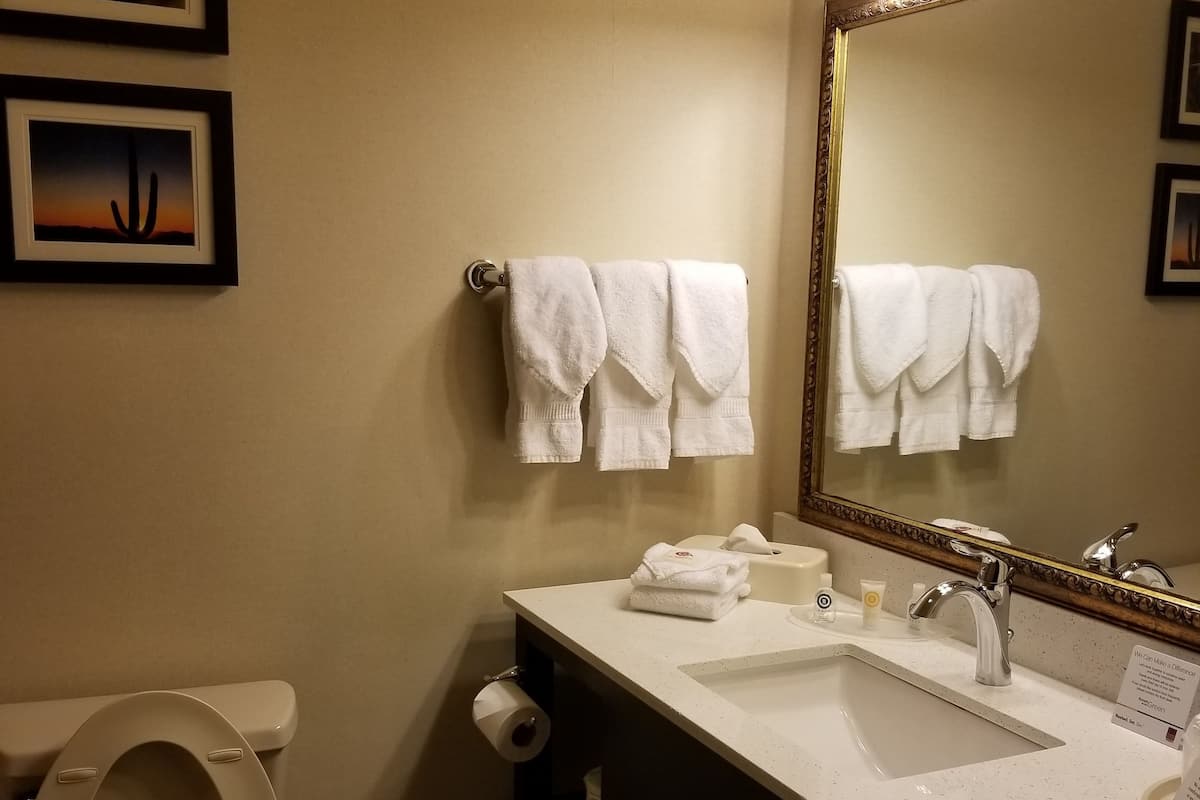 A bathroom with a toilet, sink, and mirror. There are towels hanging on the wall and a trash can.