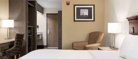 A hotel room with a bed, a desk with a chair, a bedside table with a lamp, a wardrobe, and a framed picture on the wall.