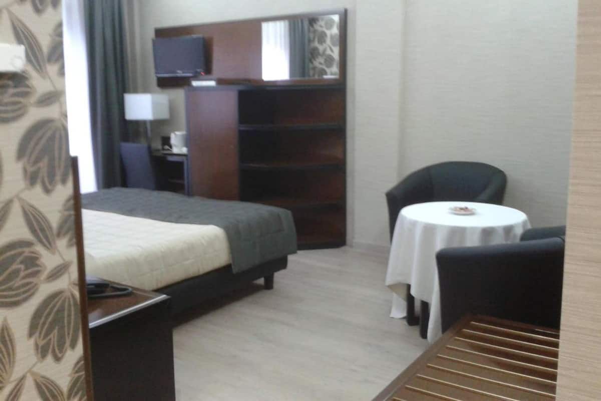 A hotel room with a bed, a desk with a lamp, a television, a bookshelf, a chair, and a small table with a tablecloth.
