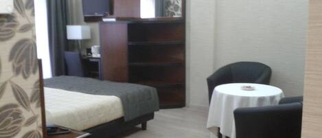 Una habitación de hotel con una cama, un escritorio con lámpara, televisión, estantería, una silla y una mesita con mantel.