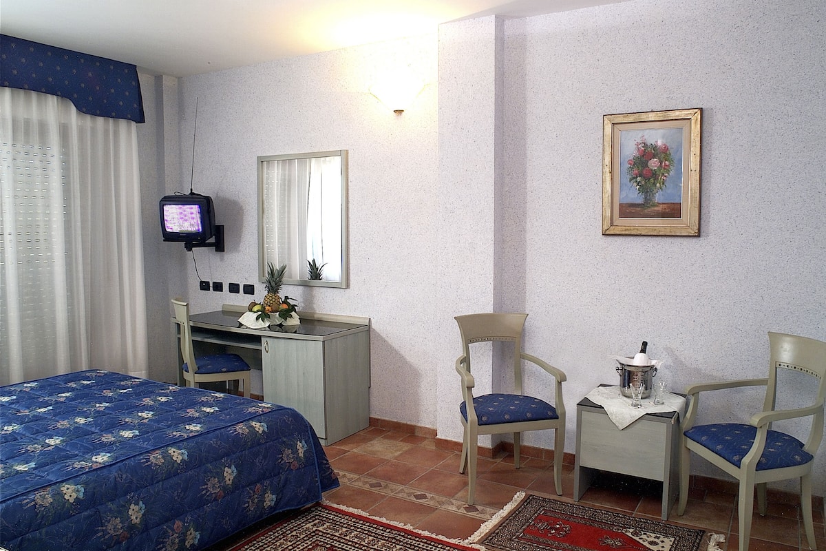 A hotel room with a bed, a desk with a computer, two chairs, a small table with a bottle and glasses, a framed picture on the wall, and a television on the wall.
