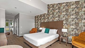 A hotel room with a large bed, two side tables, a desk, and a chair. There is a patterned wallpaper on the wall and a view of the outdoors through the door.