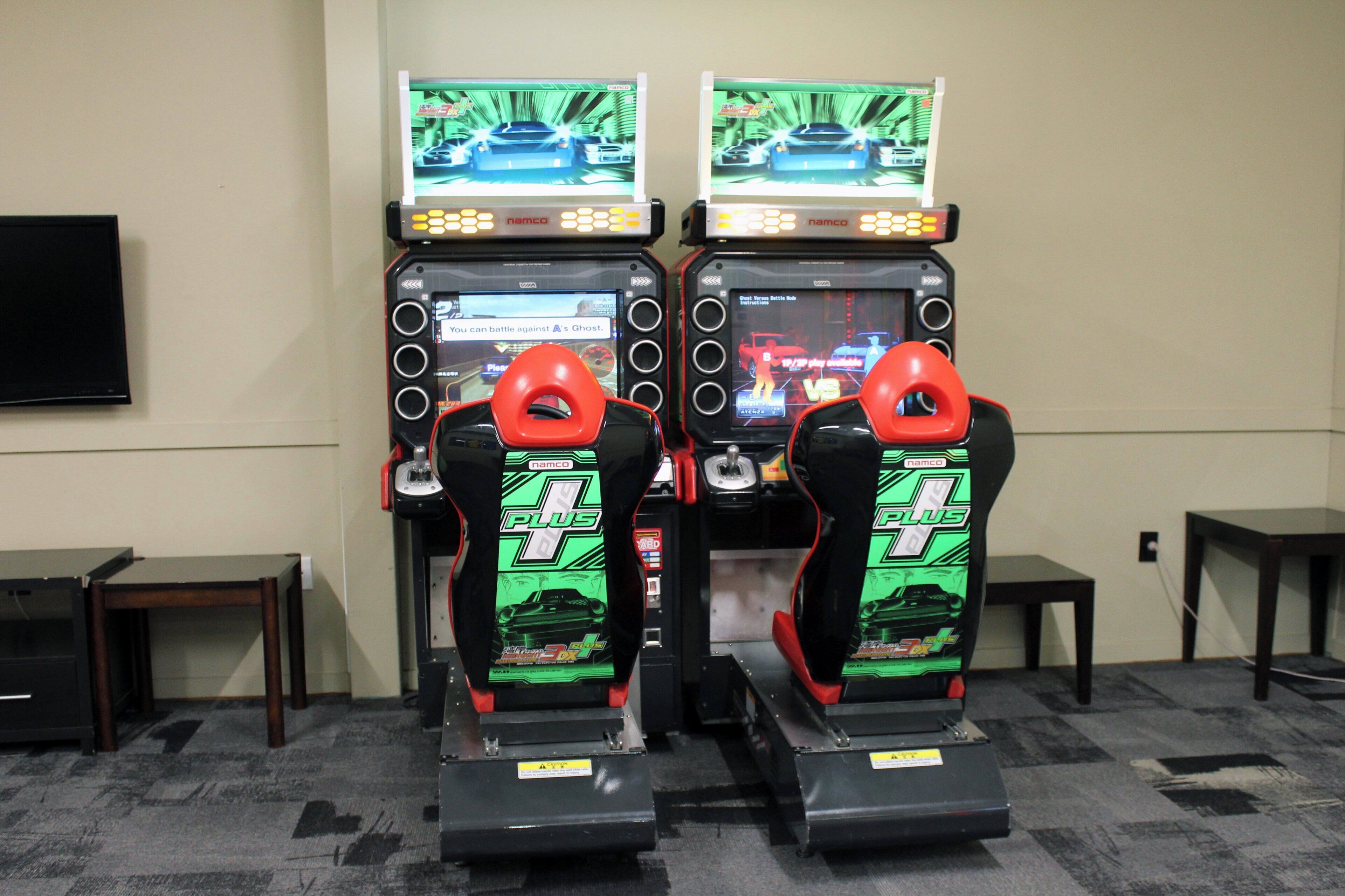 Two racing arcade machines with screens, placed in a room with carpeted flooring and a television on the wall.