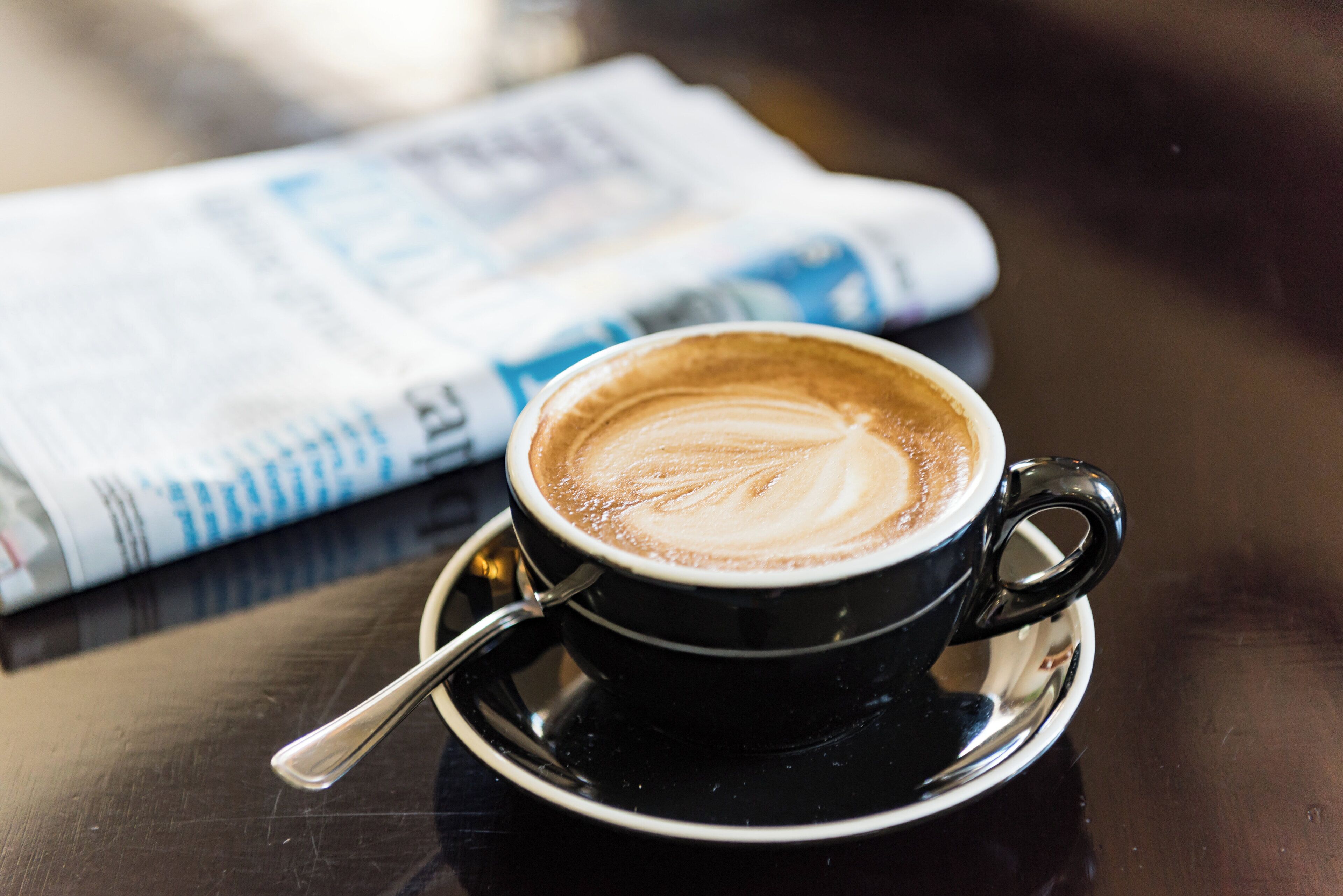 A cup of coffee with latte art on a saucer, placed on a table with a newspaper in the background.
