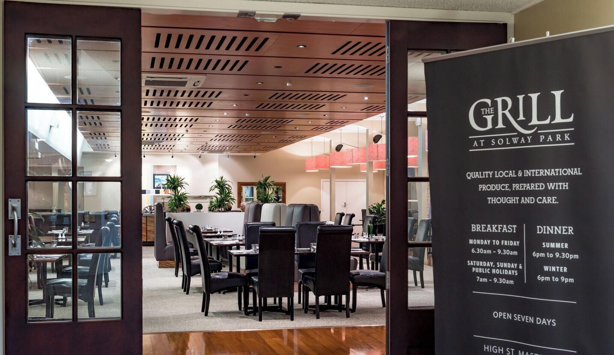 A restaurant interior with a modern design, featuring a wooden ceiling, black chairs, and a menu board advertising breakfast and dinner times.