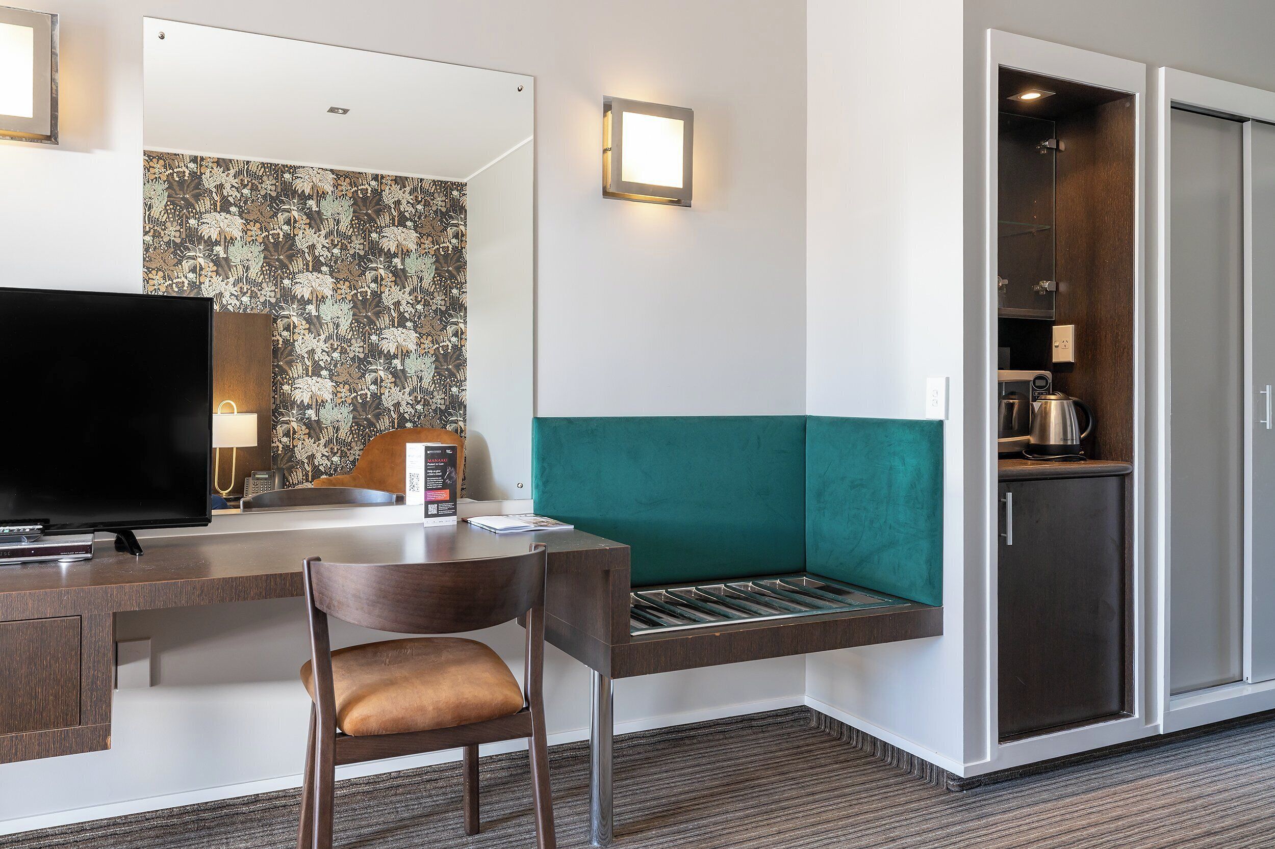 A hotel room with a TV, a desk with a chair, a bench, a mirror, a wall with a floral pattern, a light fixture, and a kitchenette.
