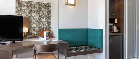 Habitación de hotel con televisión, escritorio con silla, banca, espejo, pared con estampado floral, lámpara y kitchenette.