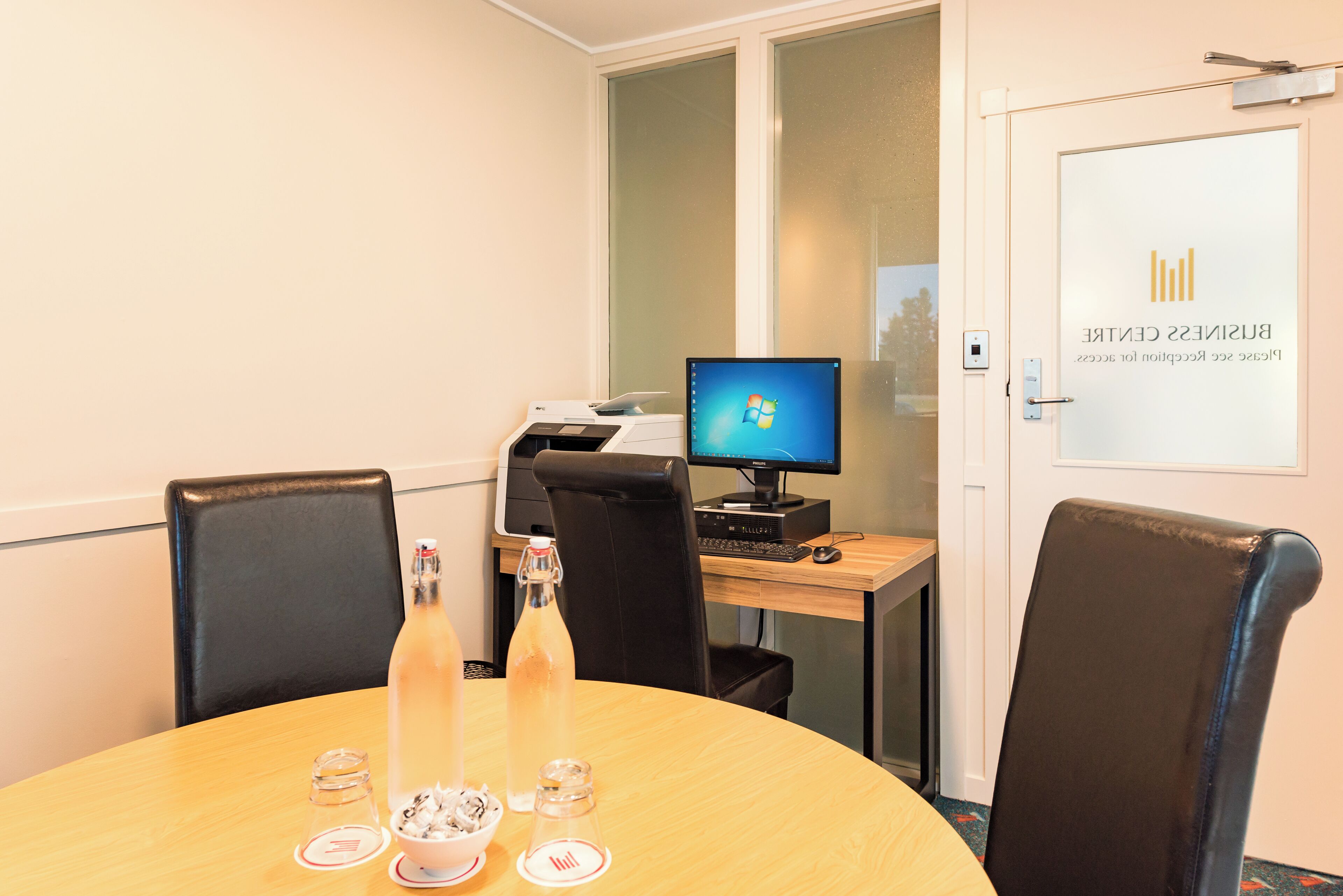 A conference room with a round table, two black leather chairs, a computer on a desk, and a door with a glass panel.