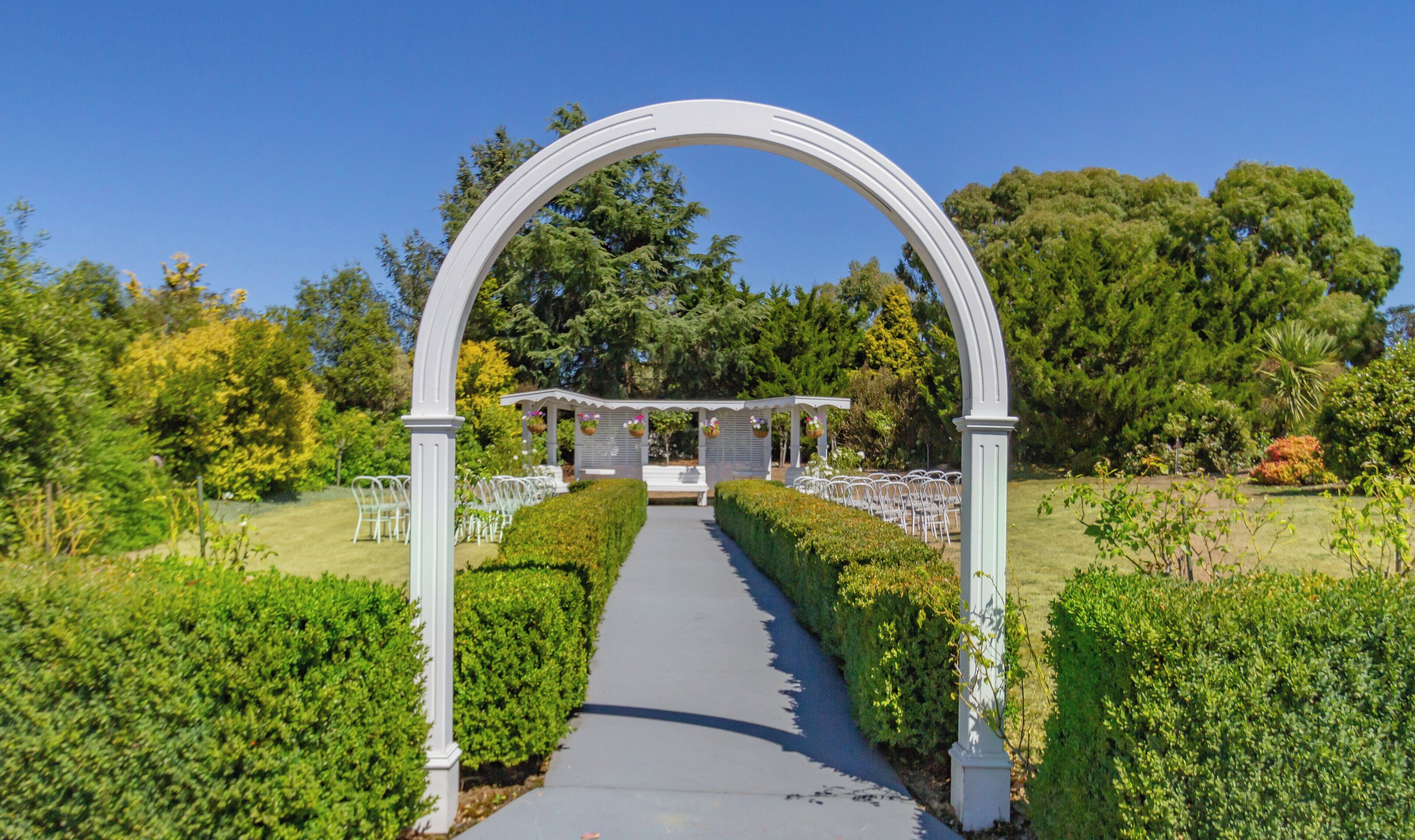 Un arco nupcial al aire libre con estructura blanca, un sendero que lleva a un pabellón y sillas ordenadas.