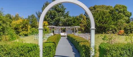 Un arco nupcial al aire libre con estructura blanca, un sendero que lleva a un pabellón y sillas ordenadas.
