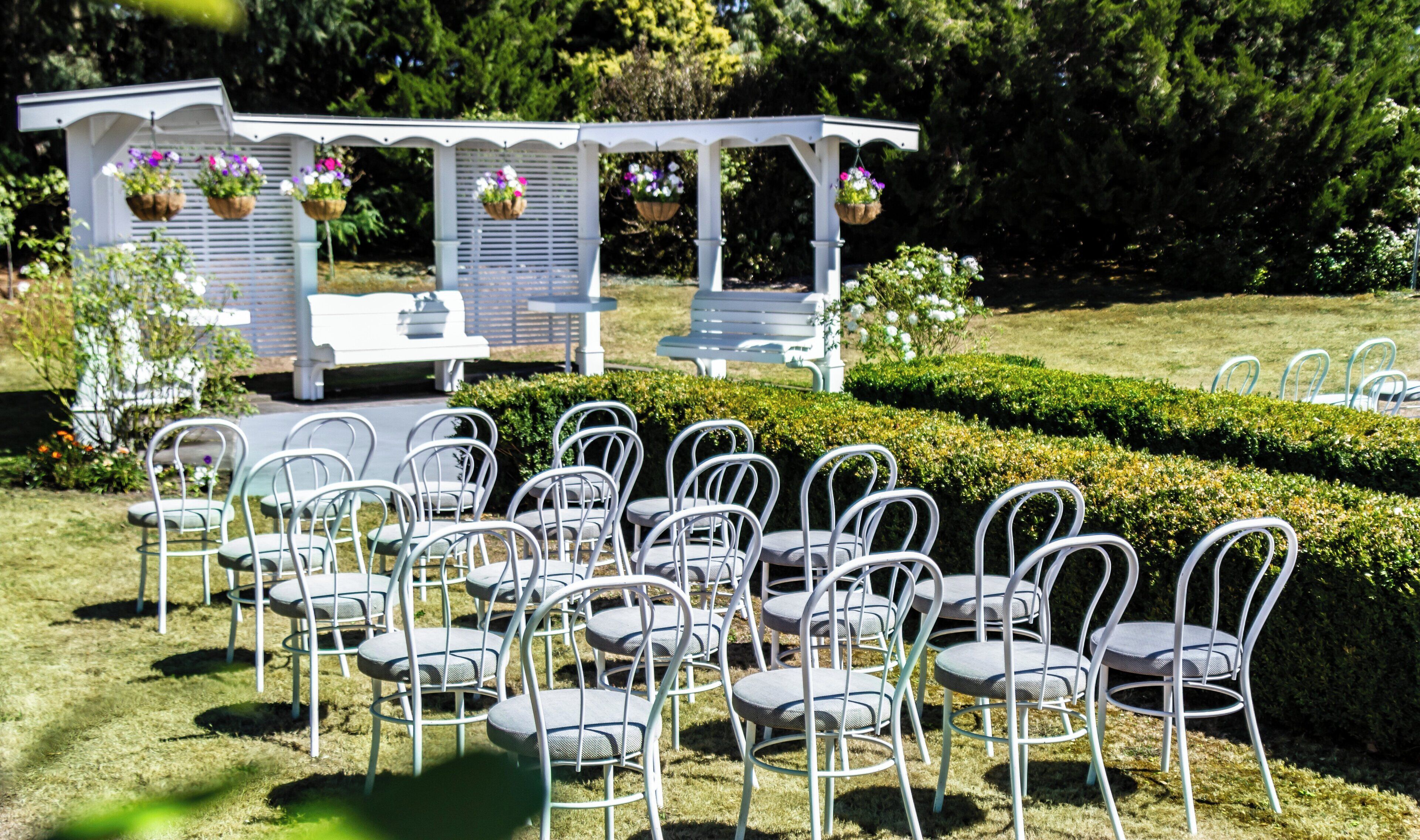 A garden setting with white chairs arranged in rows, a white gazebo with hanging flower baskets, and a hedge in the background.