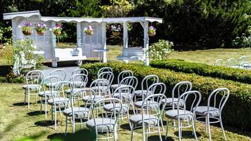 Een tuinsetting met witte stoelen in rijen, een witte pergola met bloeiende manden en een haag op de achtergrond.