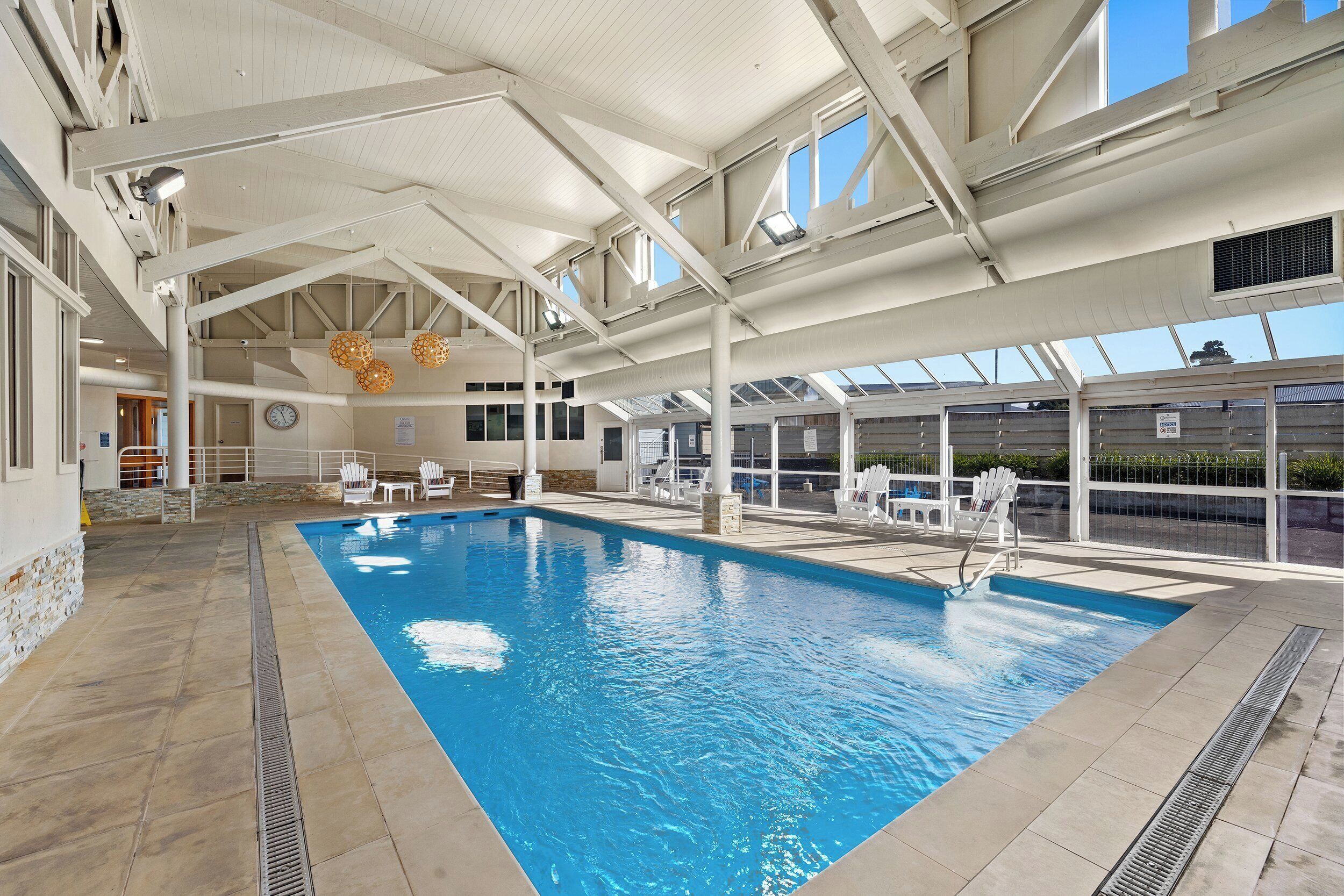 An indoor swimming pool with clear blue water, white tiled floor, and a high ceiling with visible structural beams.