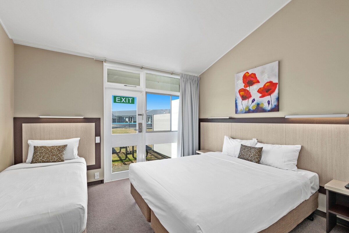 A hotel room with two beds, a painting of red flowers, and an exit sign.