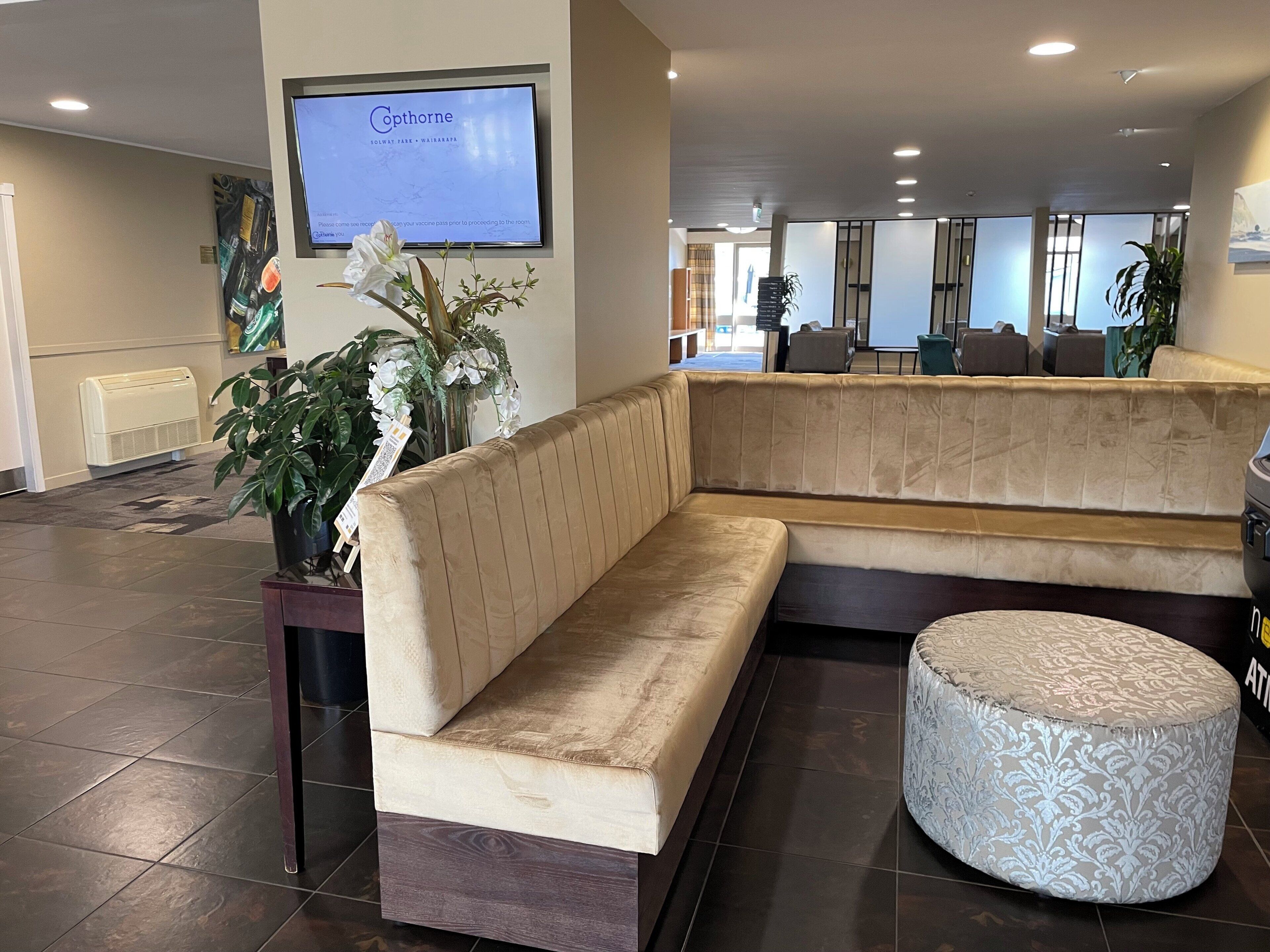 Een receptiegedeelte met een beige bank, een klein rond voetenbankje en een potplant. Een televisiescherm toont een bericht.