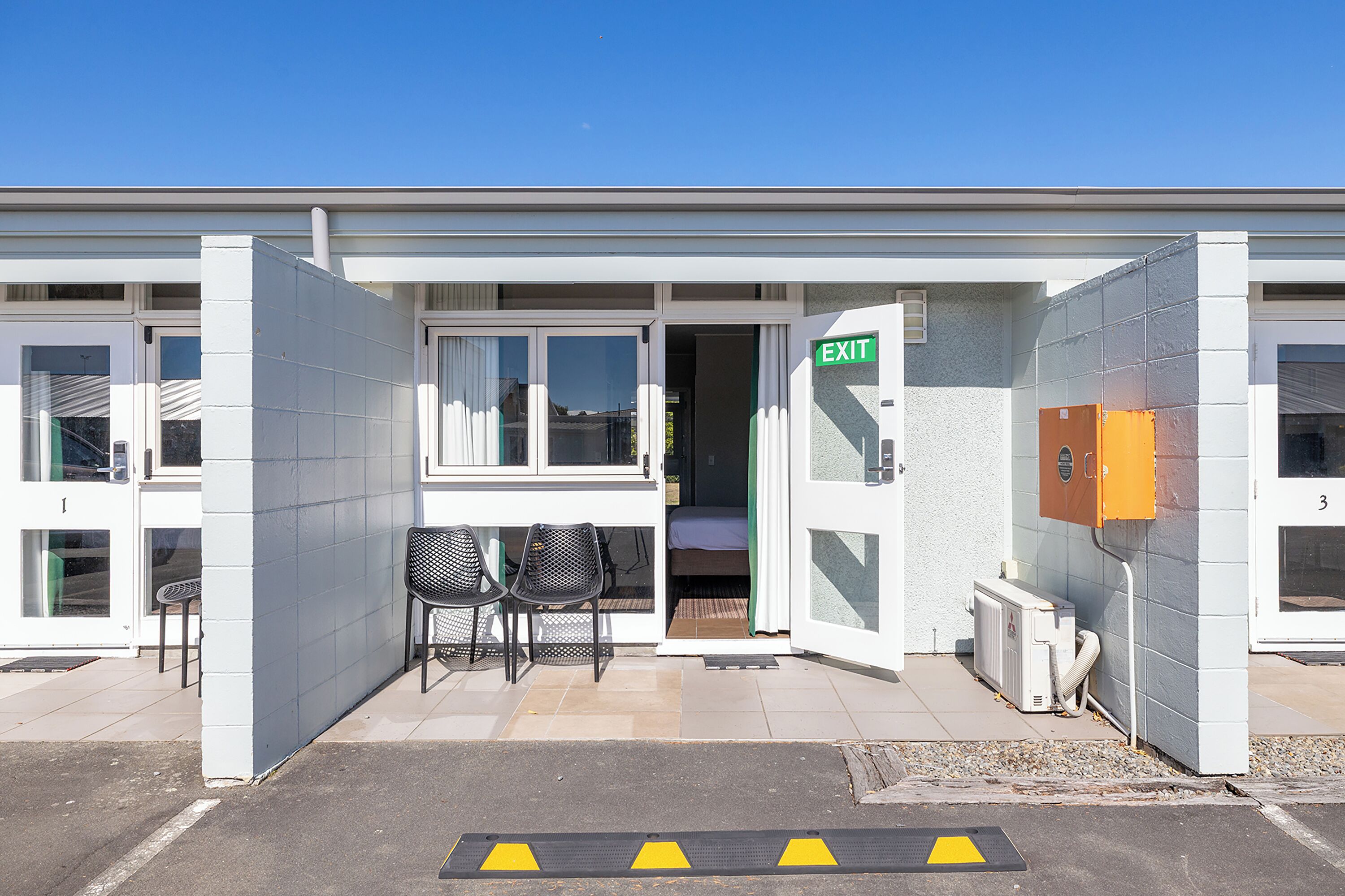 A building with an exit sign, a covered outdoor seating area with chairs, and a door leading inside.