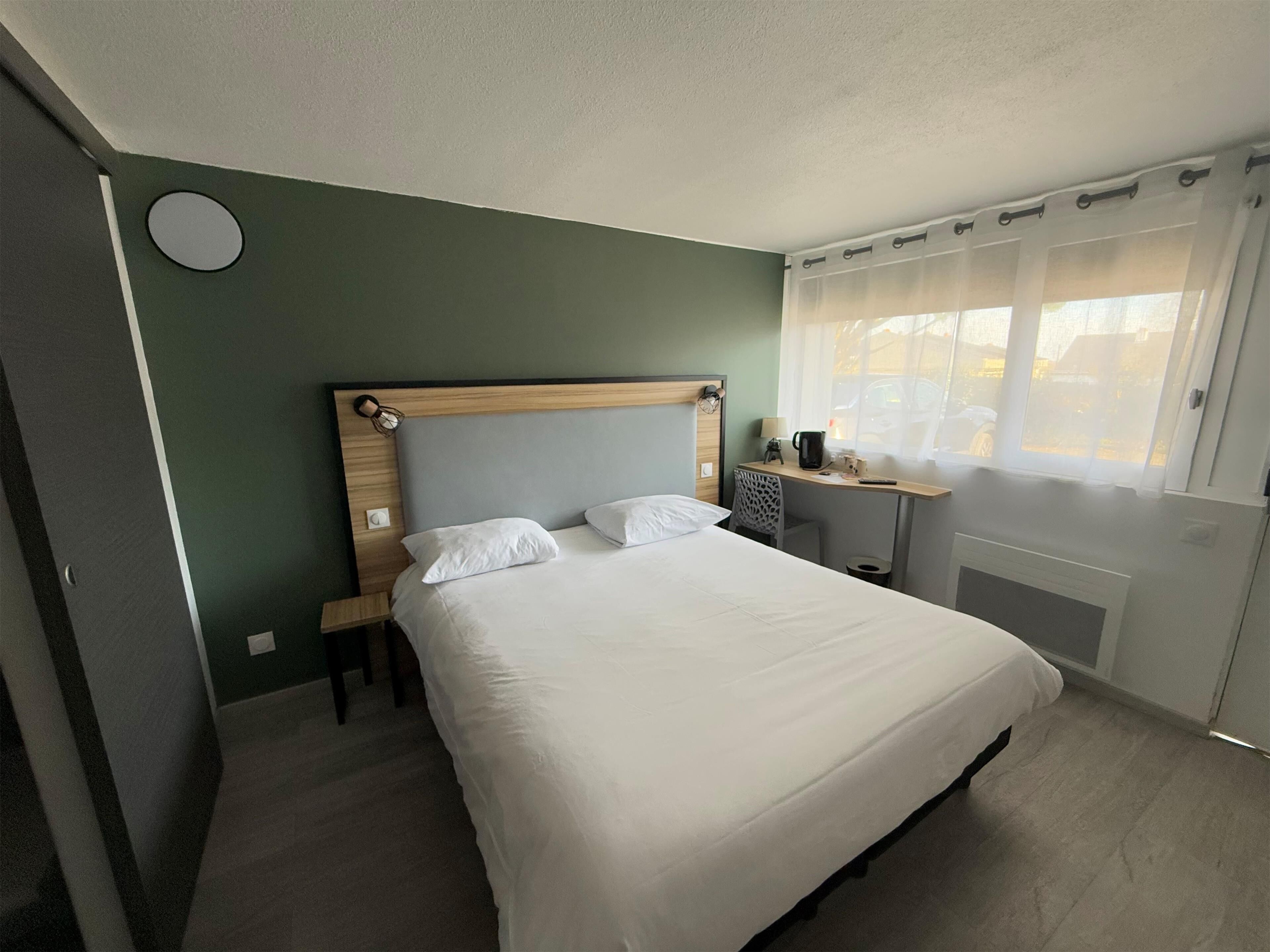 A neatly arranged hotel room with a large bed, a small desk with a chair, a window with sheer curtains, and a wall clock.